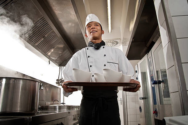 шеф-повара в белое пальто нести сковороду с чаши. - chef trainee cooking teenager стоковые фото и изображения