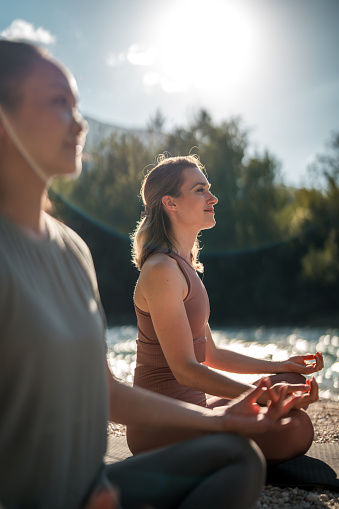 Caucasian and Asian female enthusiasts sit on yoga mats, focusing on breathing exercises and meditation, enjoying the sun by the tranquil waters of the Soča river.