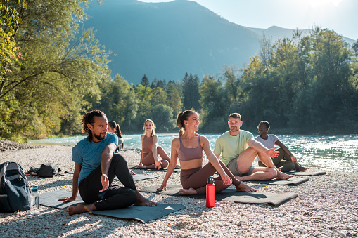 A diverse gathering practices outdoor yoga and stretching on mats by the peaceful riverbank, showcasing a mix of ethnicities.