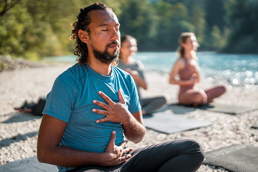 A mid adult Hispanic male sits in meditation on a yoga mat by the river.