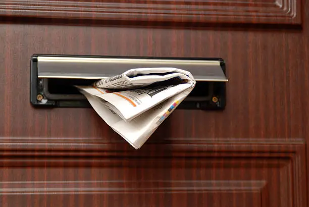 A delivered newspaper sticking out of a letter box. Focus is mainly on the newspaper.