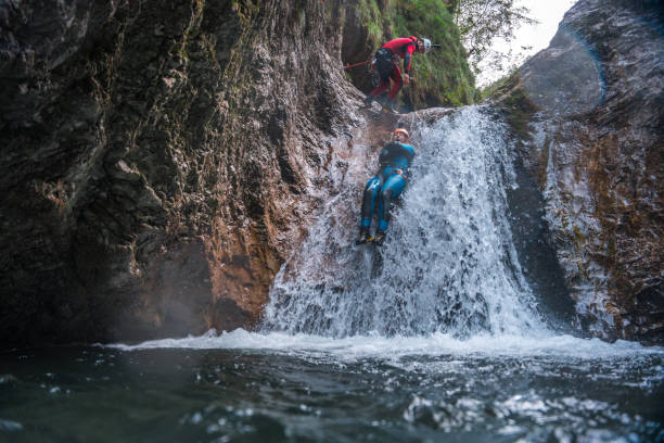 Outdoor fun, a Caucasian male, geared up in a neoprene suit and safety helmet, slides down rocky paths with the assistance of a Caucasian male guide.