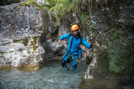 A black male, dressed in a neoprene suit and safety helmet, has a blast walking in the water during a canyoning adventure.