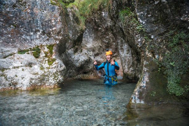 A Caucasian female in a neoprene suit and safety helmet walks through the water during a canyoning adventure.