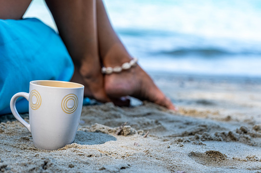Close up cup rests on the sand on the beach next to the woman's feet