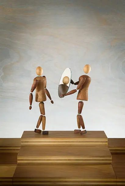 One wooden figure holds a mirror to reflect the image of a second figure. 