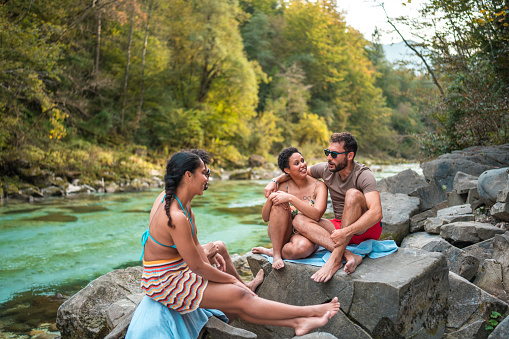 Cheerful mid adult multiracial couples delighting in a leisurely respite amid the beauty of nature. Perched on stones beside a river adorned with turquoise waters, they opt for swimsuits to combat the summer warmth.