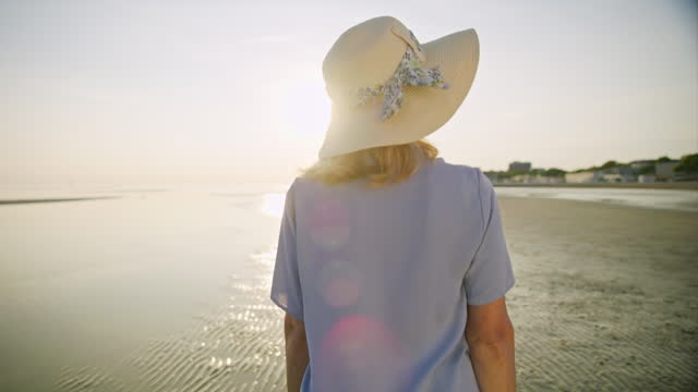 SLO MO TS Senior woman wearing a sun hat and walking on the beach at sunset