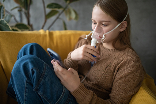 A young Caucasian girl using her smartphone while usign a nebulizer in the living room of her apartment