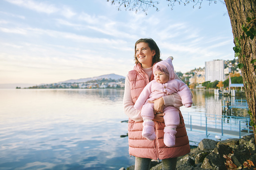 Outdoor portrait of happy young mother with adorable baby girl enjoying nice view of winter lake Geneva or Lac Leman, Montreux, Switzerland