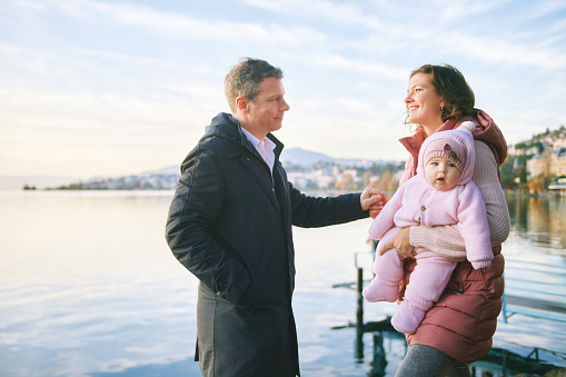 Outdoor portrait of happy young couple with adorable baby girl enjoying nice view of winter lake Geneva or Lac Leman, Montreux, Switzerland