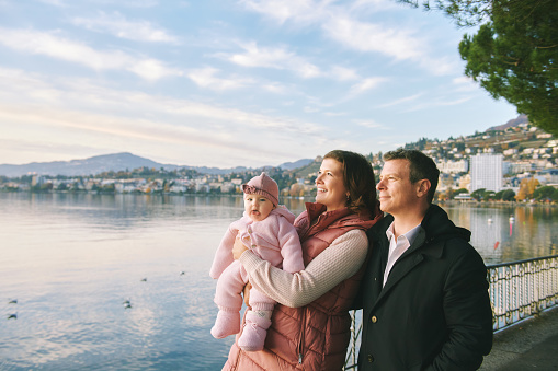 Outdoor portrait of happy young couple with adorable baby girl enjoying nice view of winter lake Geneva or Lac Leman, Montreux, Switzerland