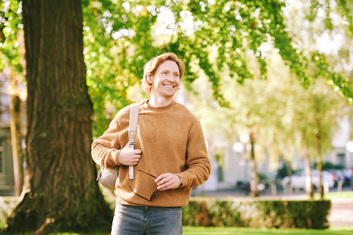 Outdoor sunny portrait of handsome happy young red-haired man in spring park, wearing brown fuzzy fleece sweater and backpack