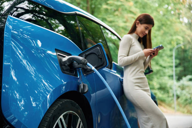 istock Leaning, using smartphone. Young woman is standing outdoors near electric automobile that is charging 1822637996