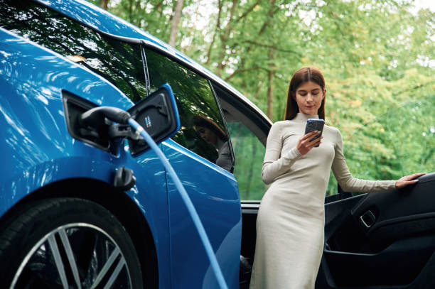 istock Leaning, using smartphone. Young woman is standing outdoors near electric automobile that is charging 1822638003