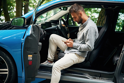 With smartphone in hands. Beautiful young man is sitting in the blue car outdoors.