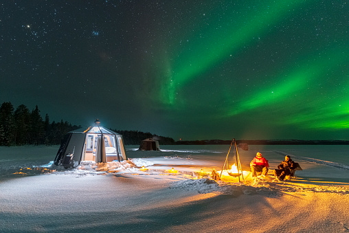 Two people on a frozen lake sitting around the fire close the glass igloo under northern lights (aurora borealis), Jokkmokk, Sweden, Lapland, Scandinavia, Europe