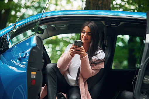 Happy beautiful woman is sitting in the blue car and using smartphone.