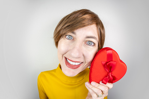 A fisheye image of a middle aged woman with short brown hair smiling as she holds a heart shaped box of chocolates for Valentine's Day.