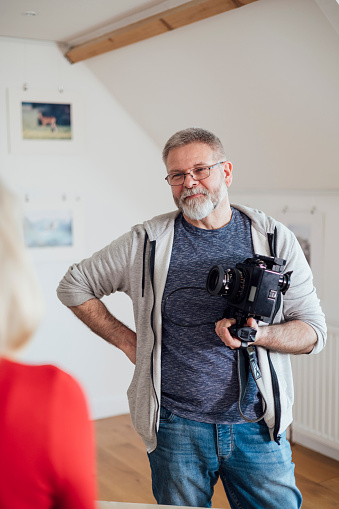 A mature man standing in a photograph gallery in Horsley, Northumberland. He is taking part in camera club and is holding a vintage camera while looking at and talking to his friend who is mostly out of shot.