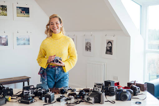 A portrait of a young woman standing in a photograph gallery in Horsley, Northumberland. She is ready to take part in a camera club and is holding a camera while looking at the camera and smiling. There are a range of different cameras on the table in front of her.