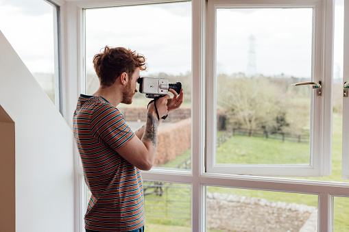A young man standing in a photograph gallery in Horsley, Northumberland. He is taking part in camera club and is holding a vintage video camera up and is filming the view outdoors through a window.