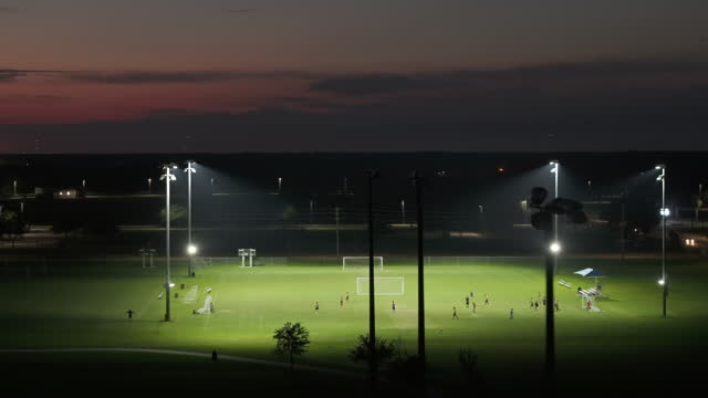 Illumination turned off on public sports arena in North Port, Florida with people playing soccer game on grass football stadium at night. Blackout concept