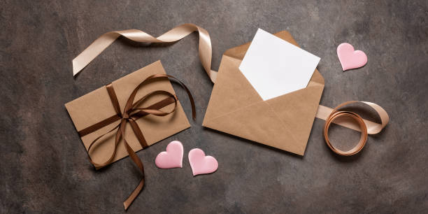 istock Open brown envelope flat lay with blank card mockup decorated with silk ribbon and silk hearts on dark vintage background. Valentine's Day card. Top view. Banner 1822690194