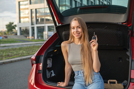 Portrait of young blonde woman with long hair sitting in the trunk of new red car and holding keys.