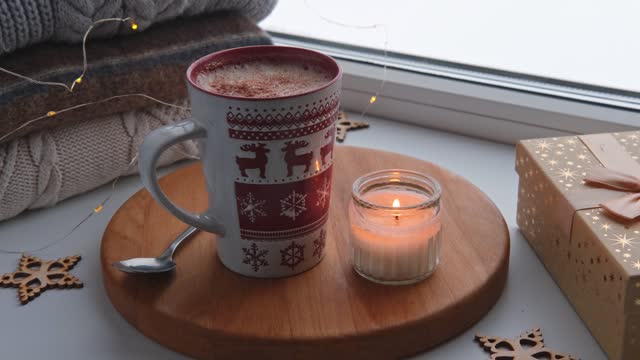 Winter windowsill still life. Red ceramic cup of hot coffee on window sill. Christmas decorations on the background. Cozy home picture. Warm woolen knitted sweaters, Burn Candle, Cookies. Stock photo