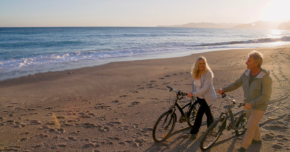Mature couple push bicycles along empty beach at sunset