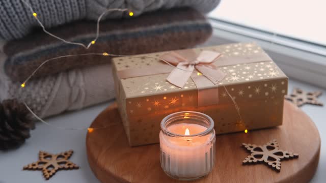Winter windowsill still life. Gift box, burning candle and cozy warm sweaters. The spirit of the Christmas holidays. Cosy home decoration concept. Warmth composition on a window sill indoors over snow