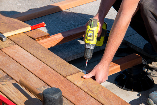 Wood deck builder building a wooden decking terrace and using power tools. Ipe wood terrace deck construction, trades skilled worker with a power screwdriver and carpenter tools installing a deck, hardwood decking