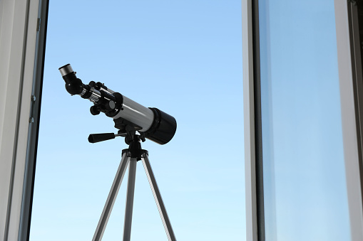 Tripod with modern telescope near open window indoors, low angle view. Space for text