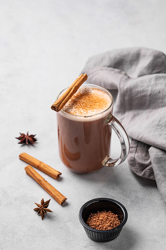 Glass mug with hot chocolate and milk foam on a light background with cinnamon sticks, anise star and cocoa powder. Warming winter  drink concept. Free space for text.