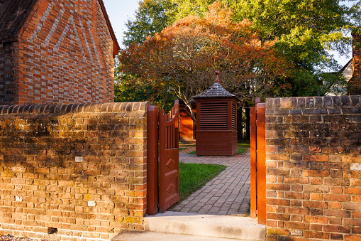 Colonial Williamsburg, Virginia, USA — October, 2023: open gate to autumnal yard with brick houses and trees in Historic District of the city. Built structure with spire stands on paving stone