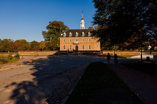 Colonial Williamsburg, Virginia, USA — October, 2023: The Capitol Building on E Duke of Gloucester, housed both Houses of the Virginia General Assembly, the Council of State and the House of Burgesses of the Colony of Virginia. People walking in the tree shadow