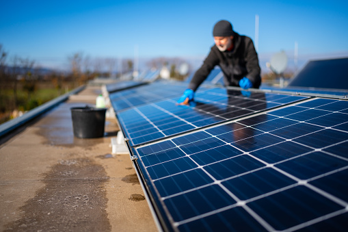 mature adult man cleaning solar and photovoltaic panels on his house roof on sunny day, focus on cells, background blurred