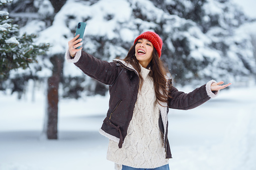 Happy woman walking in the park using her phone, taking a selfie, blogging. Young female tourist having fun in a snowy park. Concept of nature, fun, relaxation.