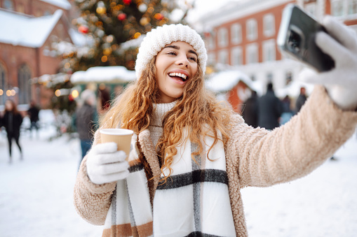 Young tourist uses a smartphone while walking through a Christmas market. Happy woman drinking hot drink and taking selfie on winter snowy day. Weekend concept.