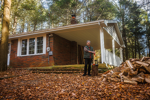 Mature Caucasian gray-haired man cutting firewood to winter. Lonely house in the forest ready in autumn season
