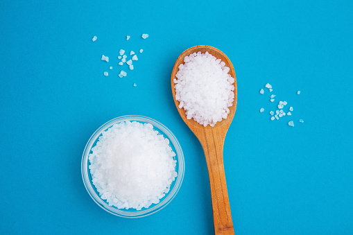 Wooden spoon with salt and glass bowl on the bright blue background. Coarse sea salt, cooking, kitchen.The benefits and harms of salt. Sodium chloride, maintaining acid-base balance in the body