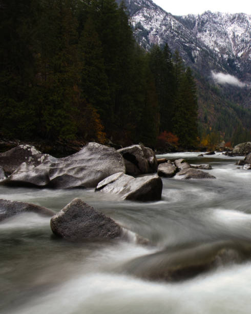 obturateur retardé avec des rapides d’eau vive lisses à travers une rivière couverte de roches dans l’état de washington avec la chaîne de montagnes north cascades en arrière-plan avec un feuillage d’automne - north cascades national park cascade range river waterfall photos et images de collection