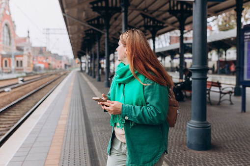 Young redhead woman waiting train with backpack and using smart phone. Railroad transport concept, Traveler. 30s Woman walking at railroad station platform.