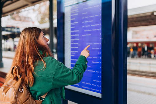Young redhead woman with backpack pointing finger on train timetable on a railway station. 30s female Looking at Arrival and Departure Information Display