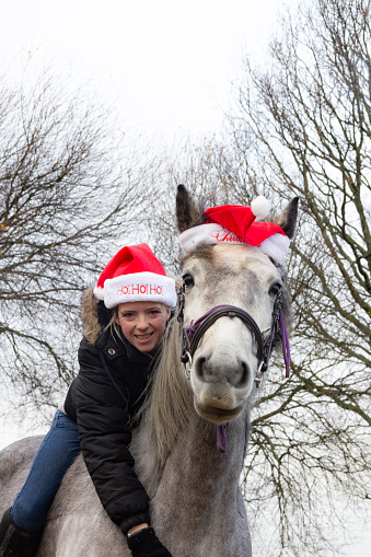 Pretty teenage girl and her grey horse both wearing red and white Santa hats, celebrating Christmas season together and having fun.