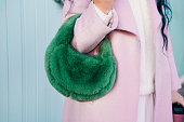 Close up cropped stylish woman in pink coat holding bright green fluffy fur bag in her hands. Fashionable modern green faux fur bag. Winter street fashion. Selective focus.