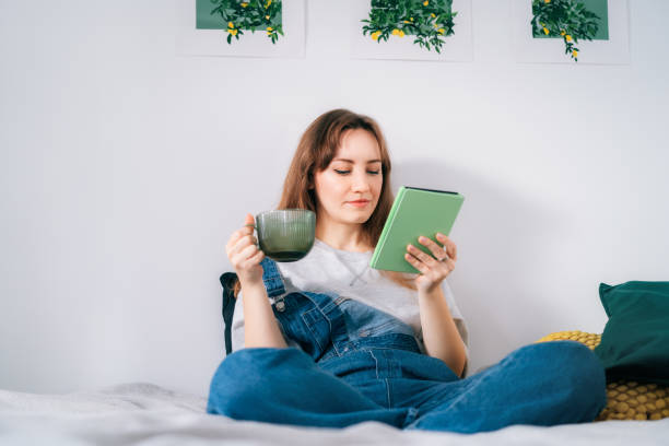 istock Young woman reading e-book on tablet reader in bed. Female relaxing with hot drink while reading an electronic book in cozy room. Mental health recovery, Personal growth. Hobby, Leisure time alone. 1823218915