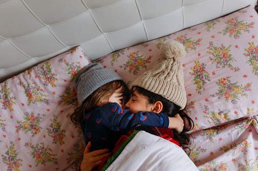 Girls wearing warm hats lay in the bed under warm blanket hug each other