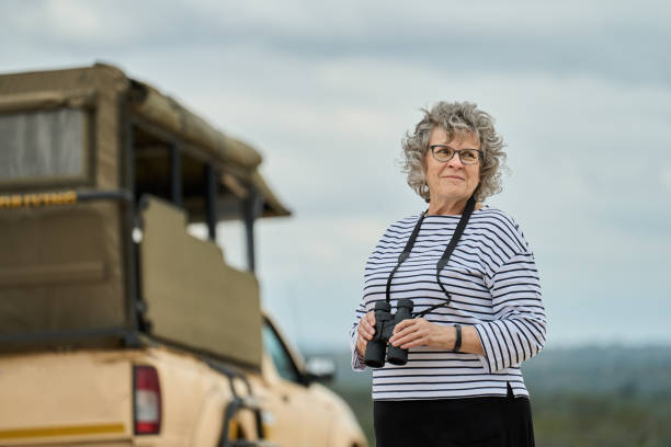 Mature woman standing with binoculars by safari vehicle on a hill overlooking the bush of a wildlife reserve